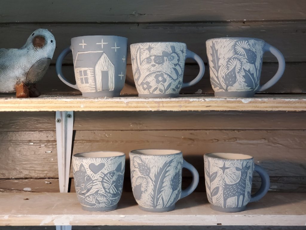 a shelf with unfired pottery, including pots decorated with a house, a dog, a goat and other animals