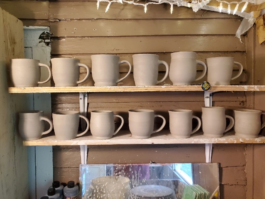 a view of the potters studio focused on 2 shelves holding newly thrown mugs