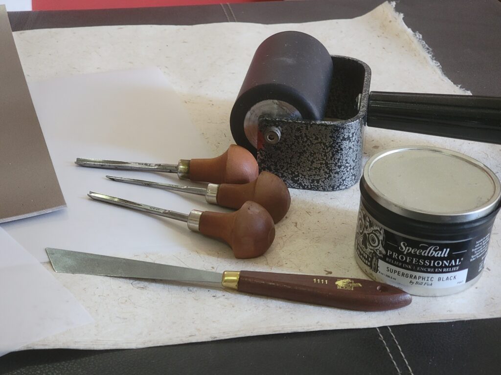photo of printmaking supplies, including paper, linoleum, brayer, knife, ink and gouges