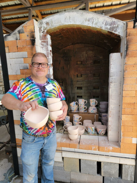artist ken swinson posing in front of a salt fired kiln with a pile of bisque pottery displayed inside it