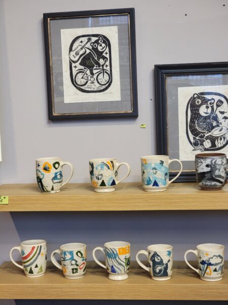 two shelves with colorful pottery cups. 2 framed linocut prints are hanging over the shelves