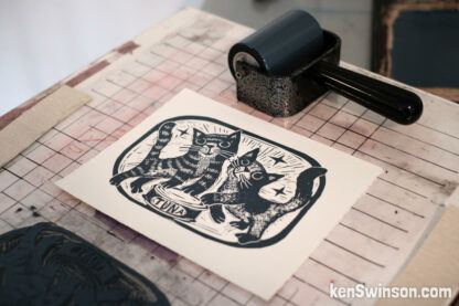 linocut depicting 2 cats looking lovingly at the viewer. a can of tuna is between them